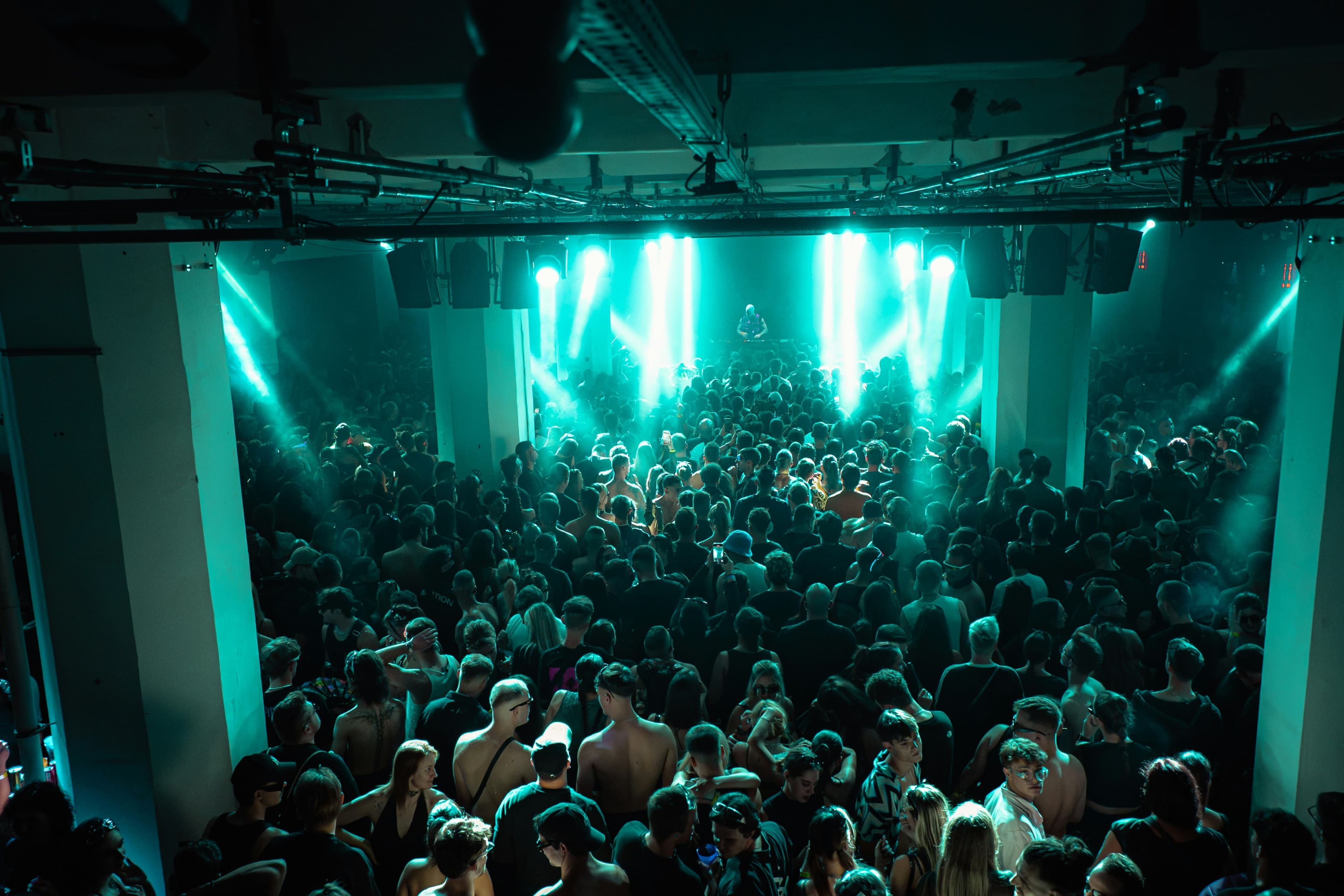 Crowded techno rave inside a warehouse venue with DJ performing under green stage lights