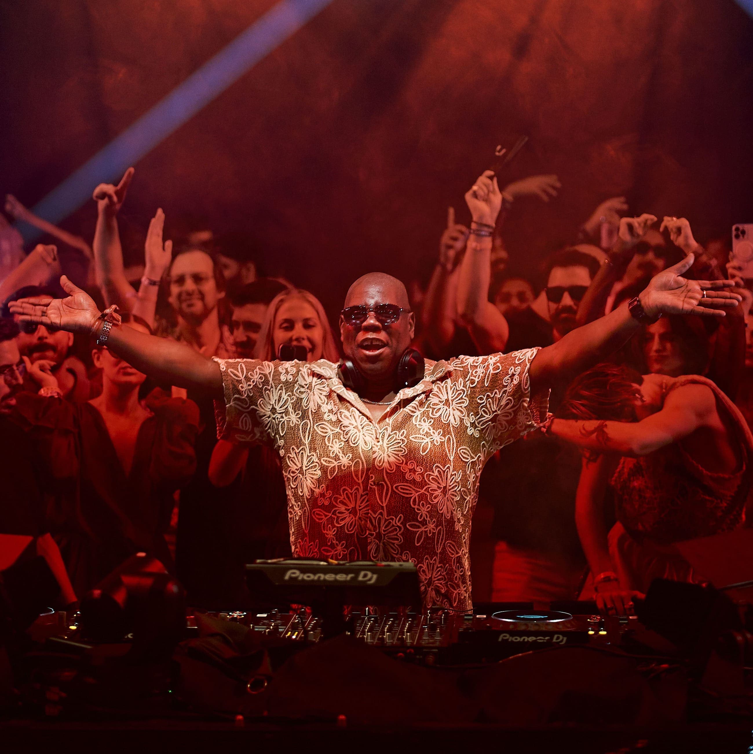 Carl Cox performing at UNVRS Ibiza, DJing in front of a packed dancefloor during his 2026 Sunday residency, with the crowd celebrating under red club lighting.